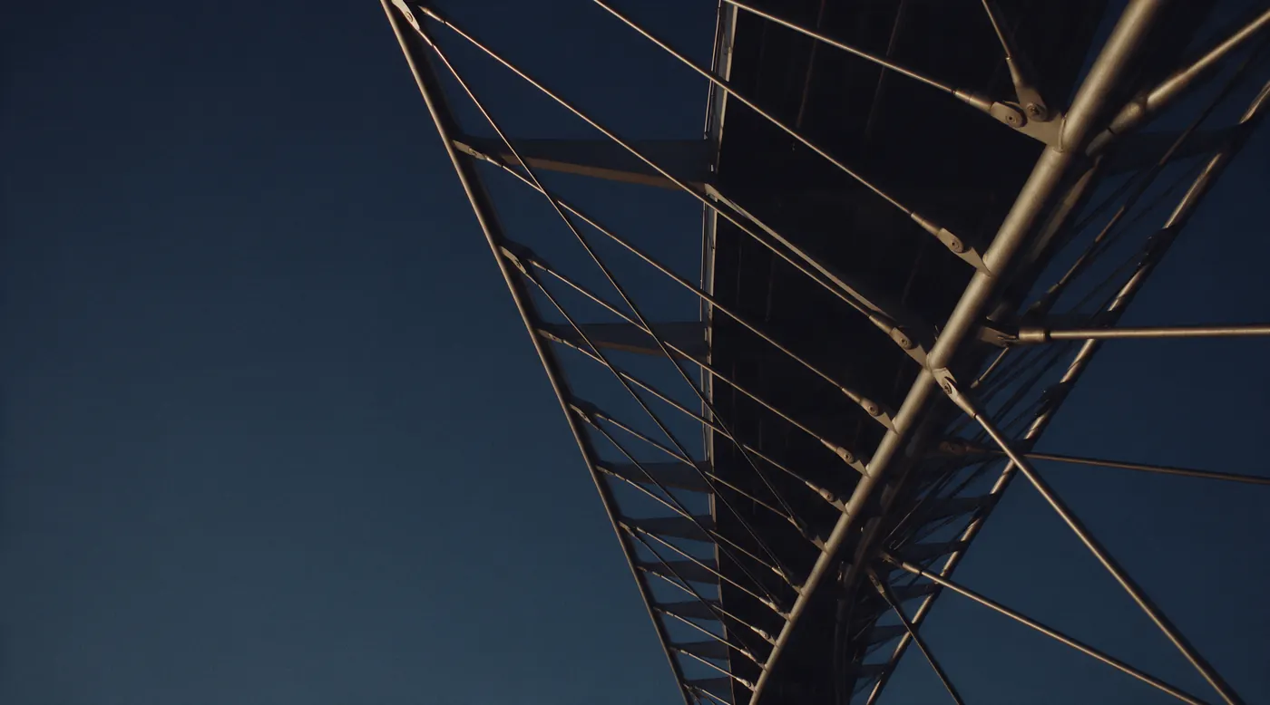 The structure of a steel footbridge seen from below at dusk, cables lit by golden light.